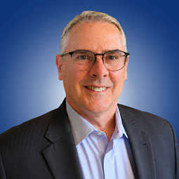 Professional headshot of a smiling man in a dark suit and light blue shirt, wearing glasses, against a blue gradient background.