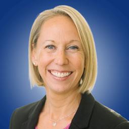 Professional headshot of a smiling blonde woman in a black blazer, pink blouse, against a blue gradient background.